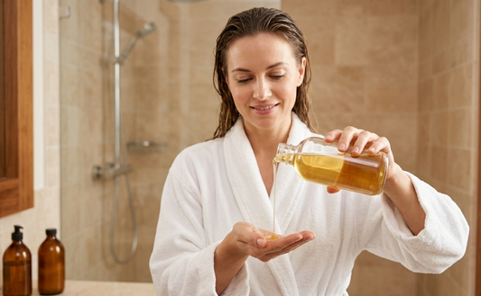 Una donna in accappatoio bianco con i capelli bagnati versa lo shampoo dorato da un flacone in vetro aperto direttamente sul palmo della mano, pronta per la detersione della cute.